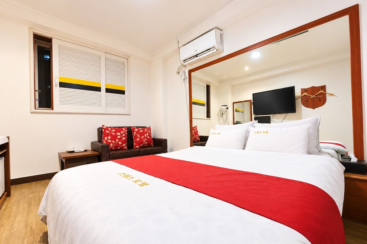 A hotel room with a large bed, a flat-screen TV, a mirror, and a window with blinds.