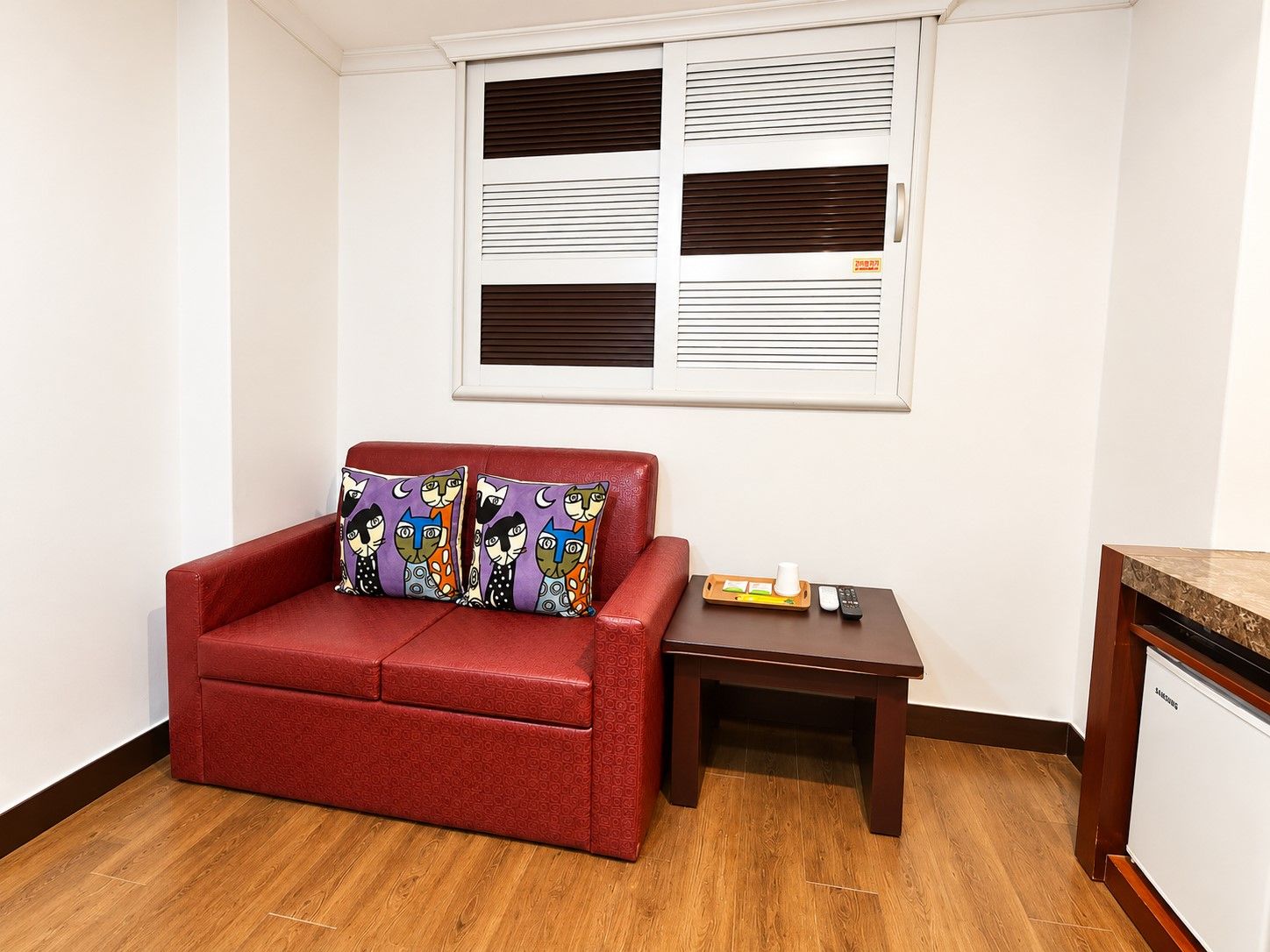 A red sofa with patterned cushions, a small wooden side table with a tray and remote, and a window with blinds.