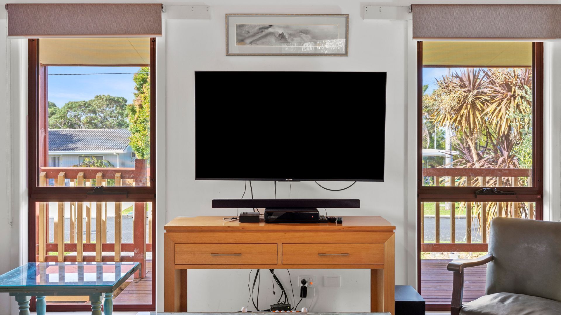 A room with a flat-screen TV mounted on the wall, a wooden desk with drawers, a chair, and large windows with a view of a residential area and trees.