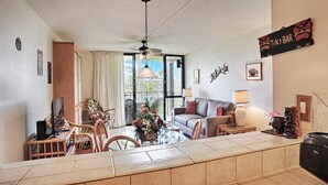 Una camera d'hotel con zona pranzo, divano, televisione e ventilatore a soffitto, con un cartello che indica 'TIKI BAR'.