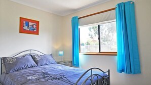 Ein Schlafzimmer mit einem Bett, einem Fenster mit blauen Vorhängen, einem Nachttisch und einem gerahmten Bild an der Wand.
