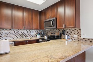 A kitchen with wooden cabinets, a stainless steel microwave, a granite countertop, and a tiled backsplash.