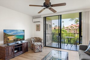 Ein Wohnzimmer mit Flachbildfernseher, Deckenventilator, Sofa, Couchtisch und einem Balkon mit Tisch und Stühlen.