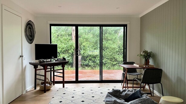 Ein modernes Wohnzimmer mit einer großen Schiebeglasdoor zu einer Holzterrasse, einem Flachbildfernseher auf einem Holzständer, einem Esstisch mit Stühlen und einer gemütlichen Sitzecke mit einem gemusterten Teppich.