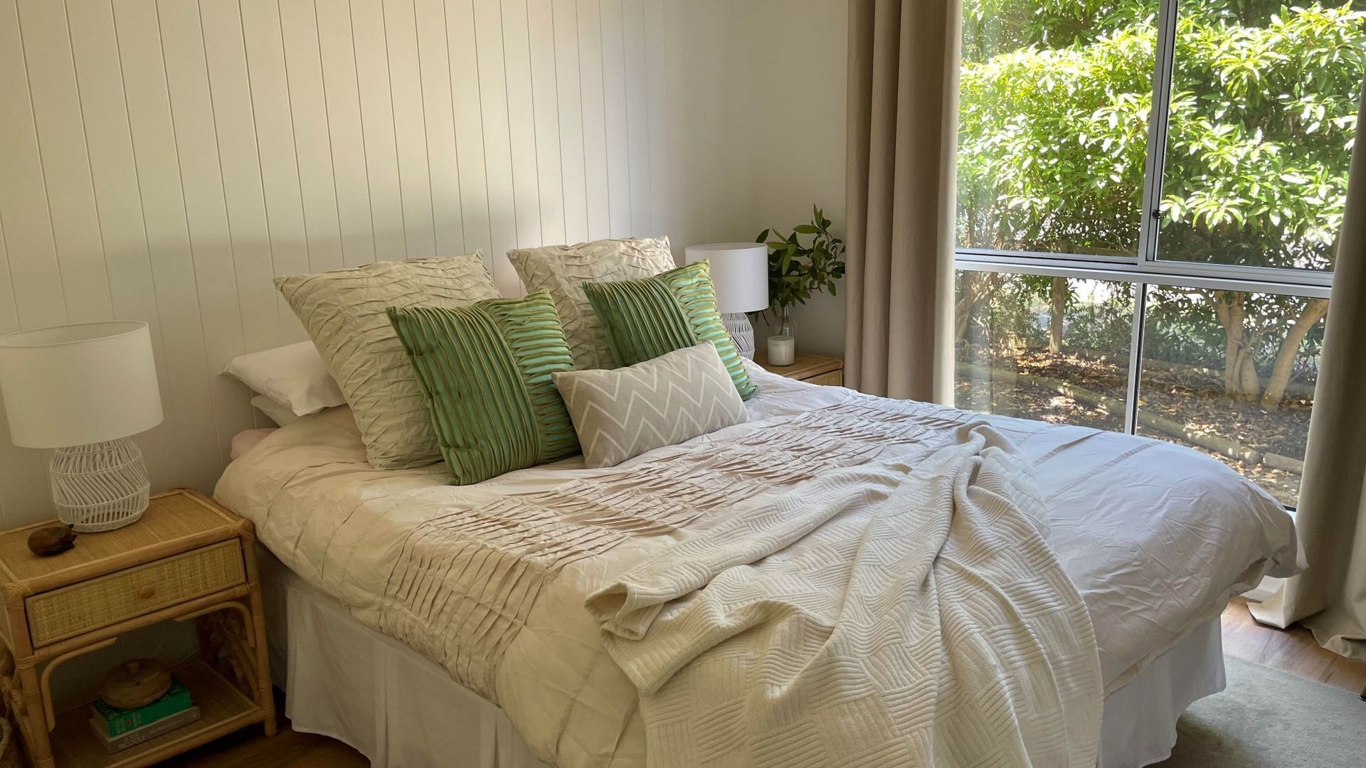 Een net opgemaakt bed met een kleurenschema van wit en beige, een bedlampje en een raam met uitzicht op groen.