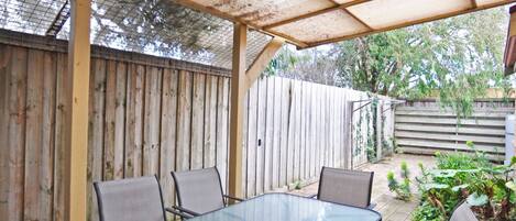 A covered patio area with a glass table and chairs, surrounded by a wooden fence and greenery.