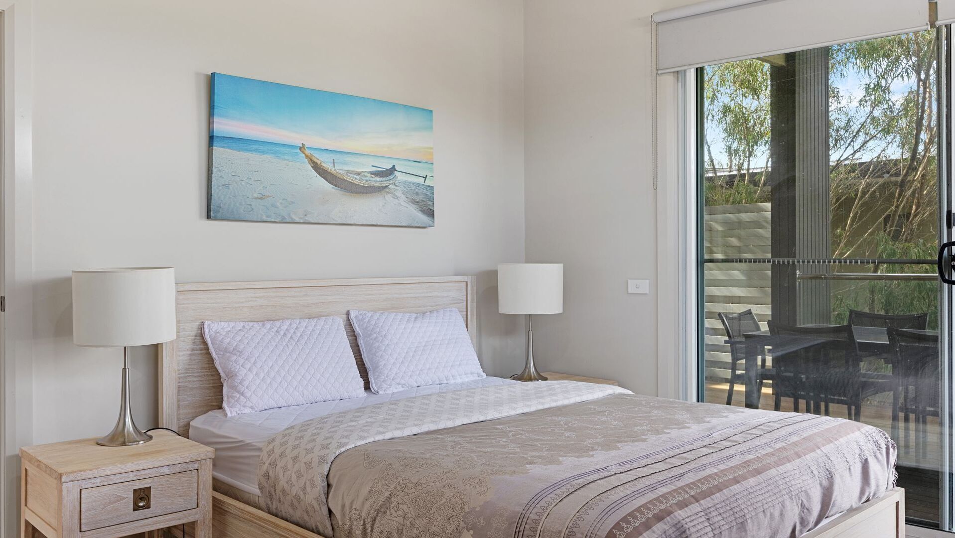 A bedroom with a bed, two bedside tables with lamps, a painting of a boat on a beach, and a sliding glass door leading to an outdoor area with chairs.