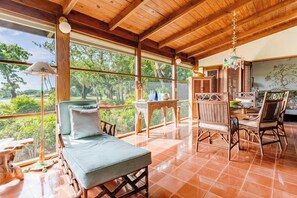 A room with a bed, a table, chairs, and large windows offering a view of greenery.