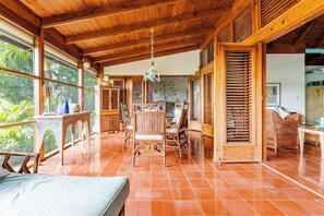 A spacious room with a wooden ceiling, a dining area with wicker furniture, and a view of greenery outside.
