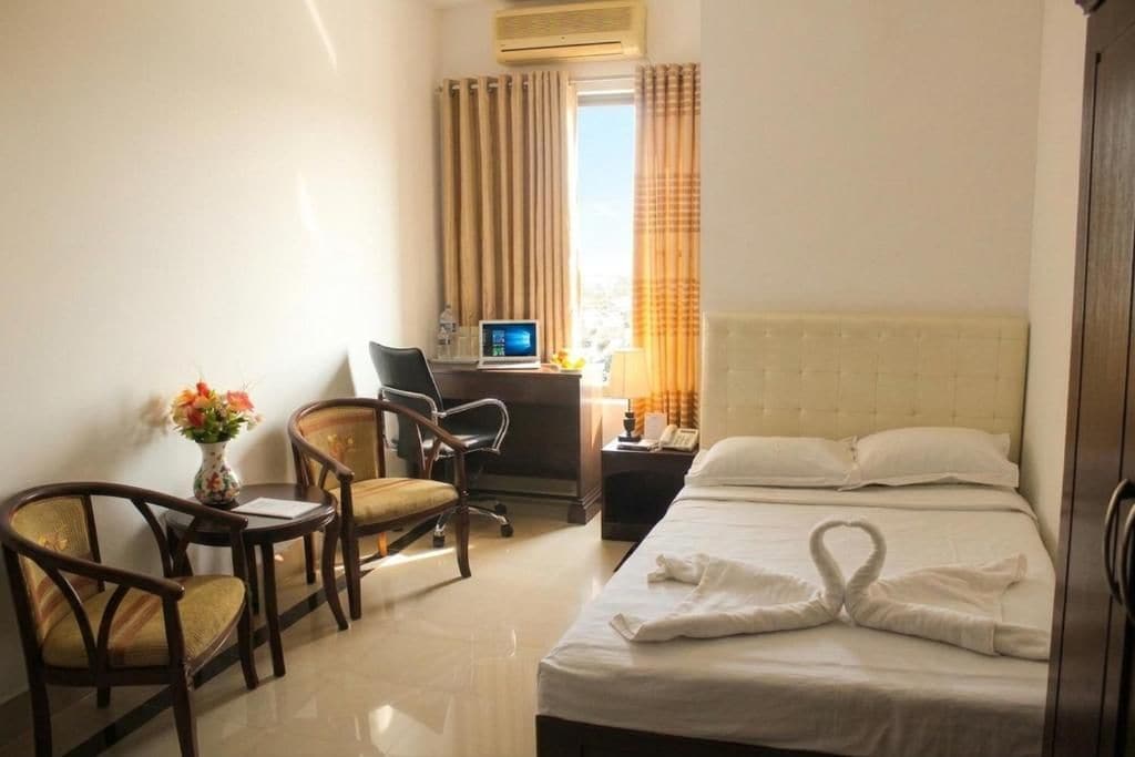 A hotel room with a bed, two chairs, a desk with a computer, a vase with flowers, and a window with curtains.