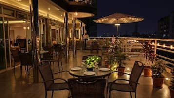 An outdoor dining area with tables and chairs, potted plants, and a view of the cityscape at night.