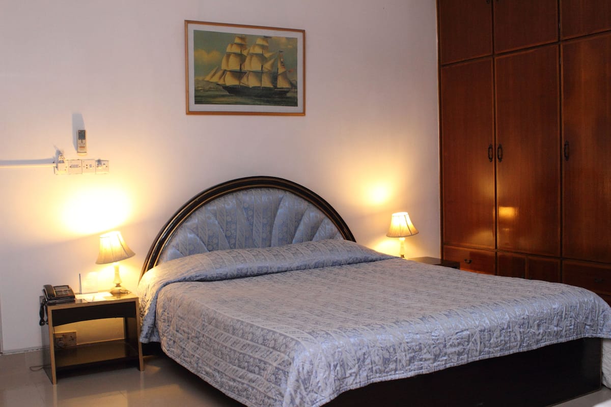 A neatly made bed with a patterned bedspread, two bedside lamps, a nightstand with a phone, and a painting of a ship on the wall.