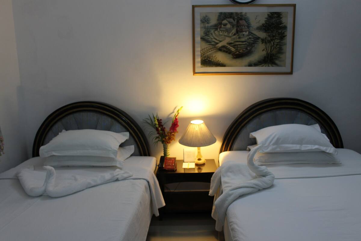 A hotel room with two single beds, a nightstand with a lamp, and a framed picture on the wall.