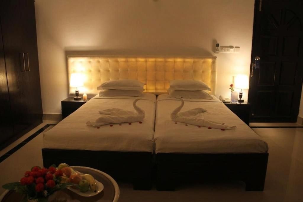 A hotel room with two beds, a headboard, bedside tables, and a small table with fruit and flowers.