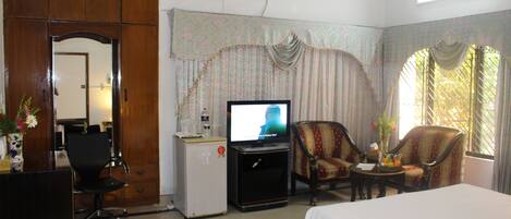 A hotel room with a bed, a desk with a computer, a television, two armchairs, and a small table with a vase of flowers.