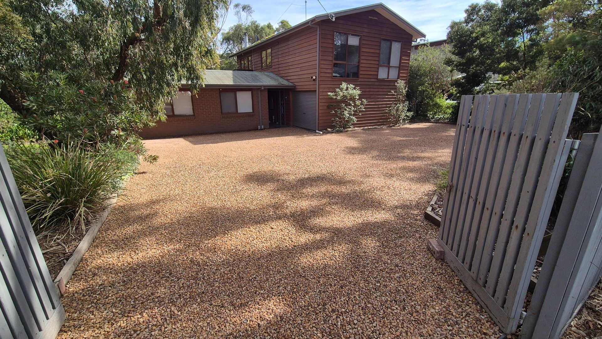 A gravel driveway leading to a wooden house with a metal gate.