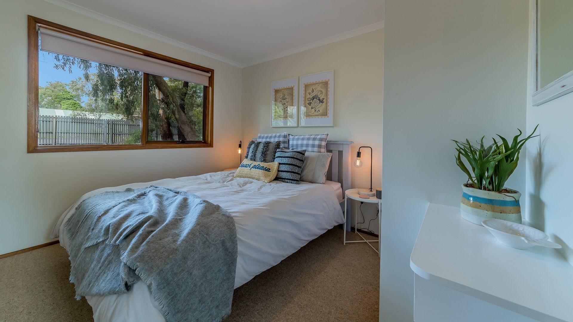 A bedroom with a bed, a bedside table, a lamp, a window with a view of trees, and a wall-mounted plant.