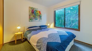 A bedroom with a large window, a bed with a patterned quilt, a small bedside table with a lamp, and a colorful painting on the wall. - Pescarina (INVERLOCH)