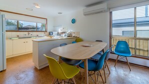 A compact kitchen with a dining area, featuring a wooden table, blue and yellow chairs, and a small island with a sink. - Pescarina (INVERLOCH)
