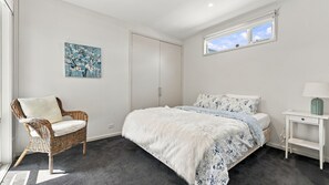 A bedroom with a large bed, a wicker chair, a nightstand, a lamp, a wardrobe, and a painting on the wall.