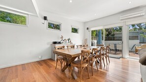 Un comedor amplio con una mesa y sillas de madera, una zona de cocina con encimera blanca y ventanas grandes que dejan entrar luz natural.