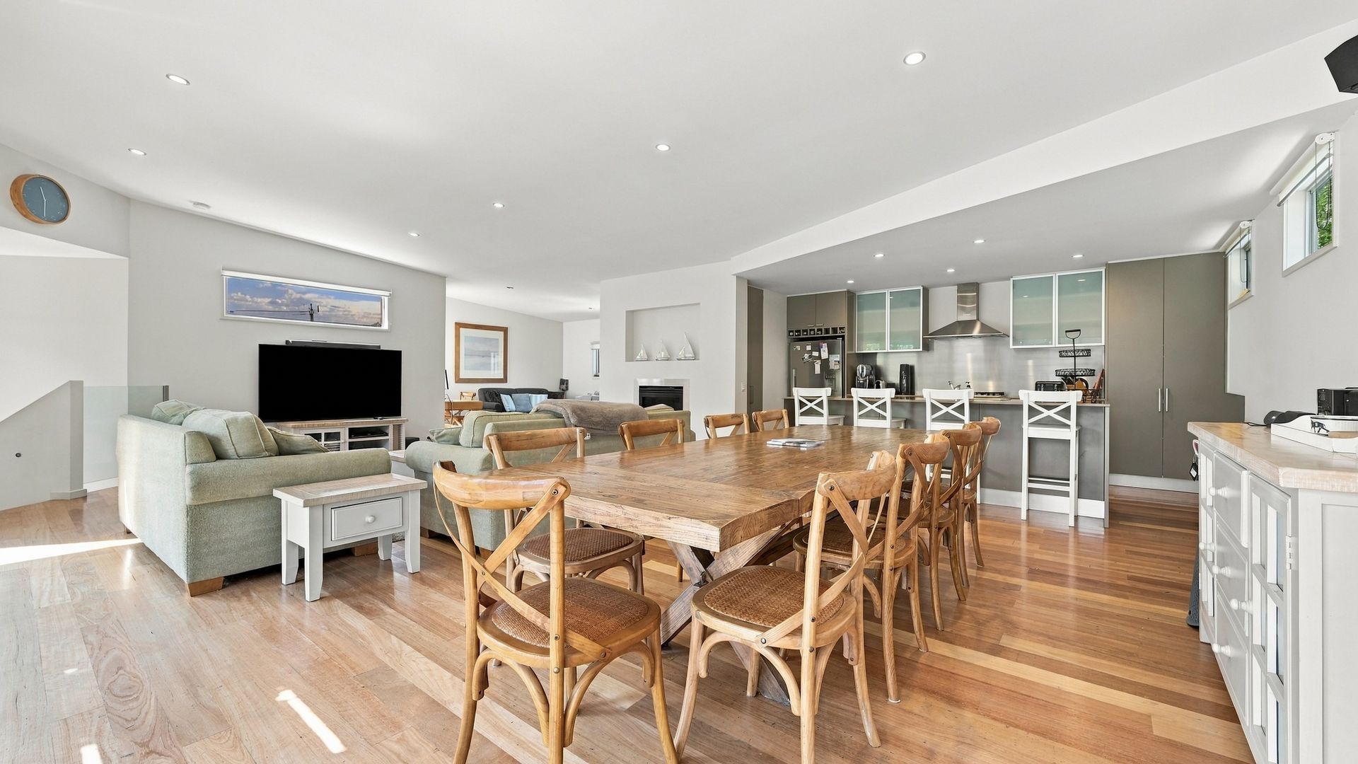A spacious living area with a large dining table, multiple chairs, a sofa, a flat-screen TV, and a kitchen area with cabinets and a stove.