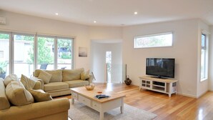 Ein modernes Wohnzimmer mit einem beige farbenen Sofa, einem Couchtisch, einem Fernseher auf einem Holzboden und großen Fenstern, die natürliches Licht hereinlassen.