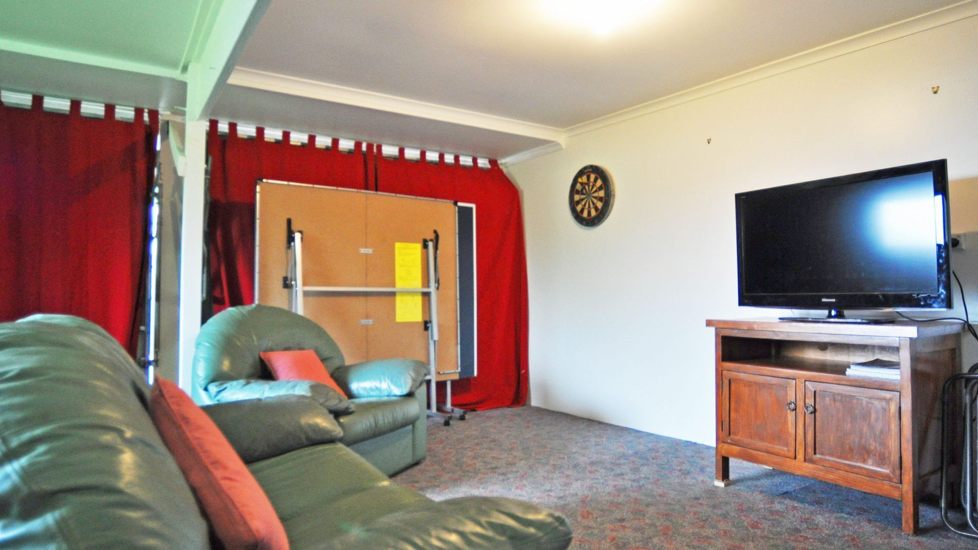 A living room with a green sofa, a television on a wooden stand, a dartboard, and a red curtain.