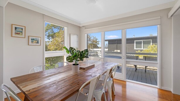A spacious room with a wooden dining table, white chairs, and large windows offering views of the outdoors.