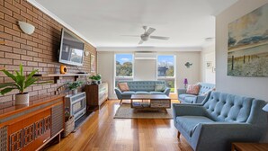 A spacious living room with a brick wall, a flat-screen TV, a ceiling fan, and a large window offering a view of the outdoors.