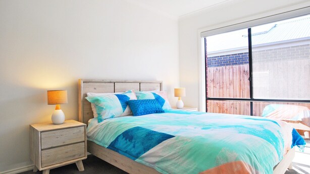 A bedroom with a bed, two bedside tables, and a window with a view of a wooden fence. - Plovers Rest Inverloch (INVERLOCH)