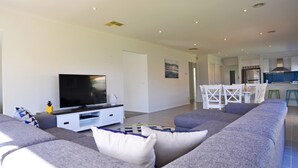 A modern living room with a large flat-screen TV on a white entertainment unit, grey sofas with patterned cushions, and a dining area with white chairs and a wooden table. - Plovers Rest Inverloch (INVERLOCH)