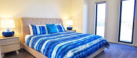 Un dormitorio con una cama grande, dos mesitas de noche con lámparas, un cabecero acolchado y vistas al exterior a través de una puerta corredera.