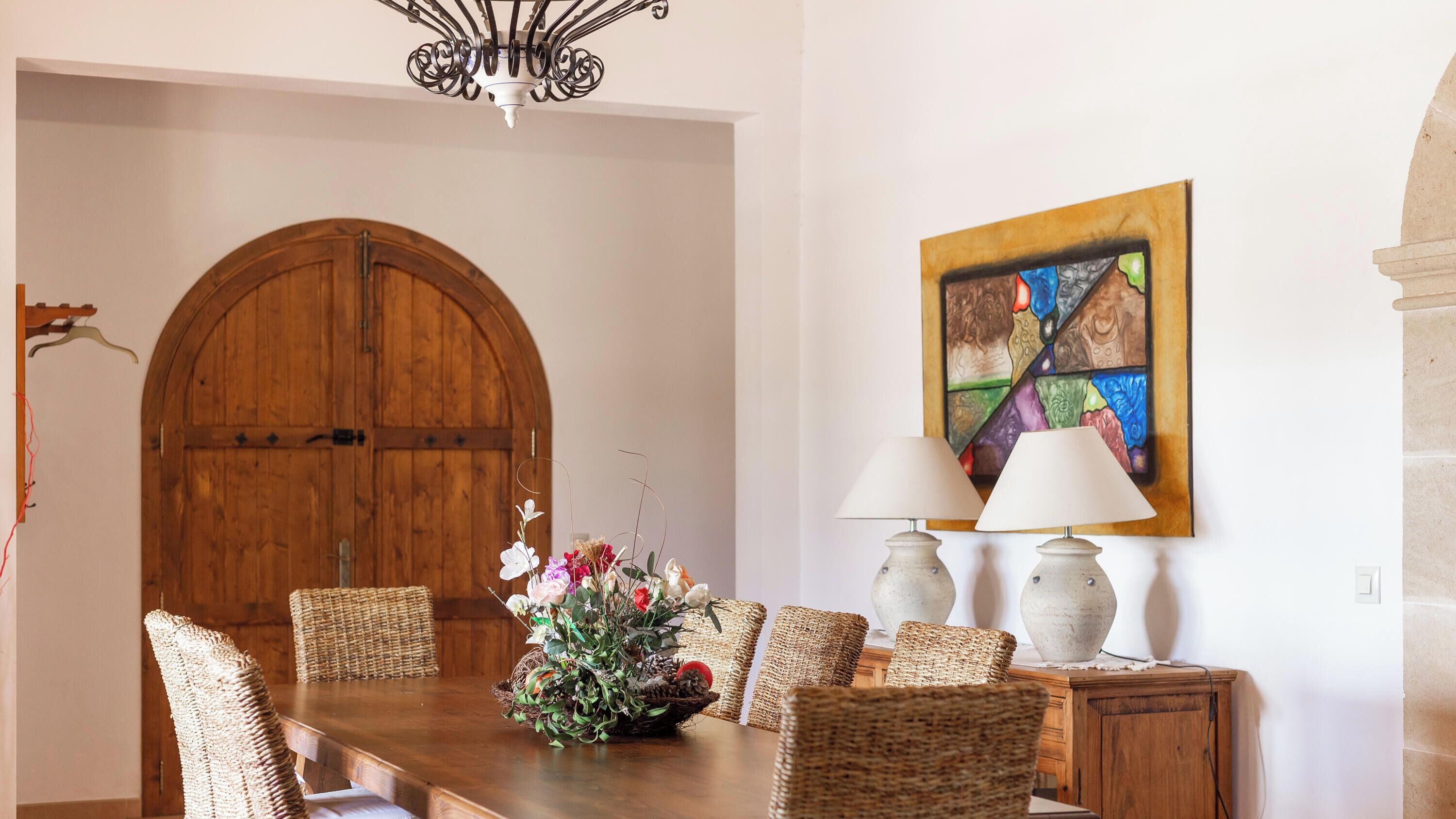 A dining area with a wooden table, wicker chairs, a floral centerpiece, and a painting on the wall.