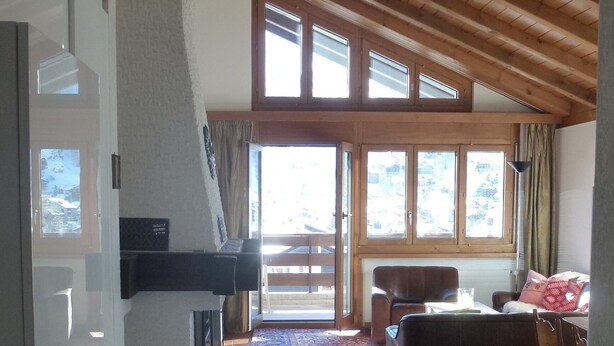A spacious living area with a high ceiling, wooden beams, a large window, a sofa, a coffee table, and a patterned rug.