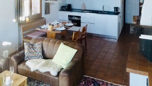 A cozy living room with a brown leather sofa, a dining table set for four, and a kitchen area with white cabinets and a wooden ceiling.