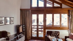 A living room with a high ceiling, wooden beams, a television, a sofa, a coffee table, and a patterned rug.