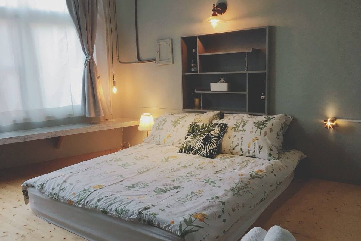 A bed with floral bedding, a bedside lamp, a shelf with a bottle, and a window with curtains.