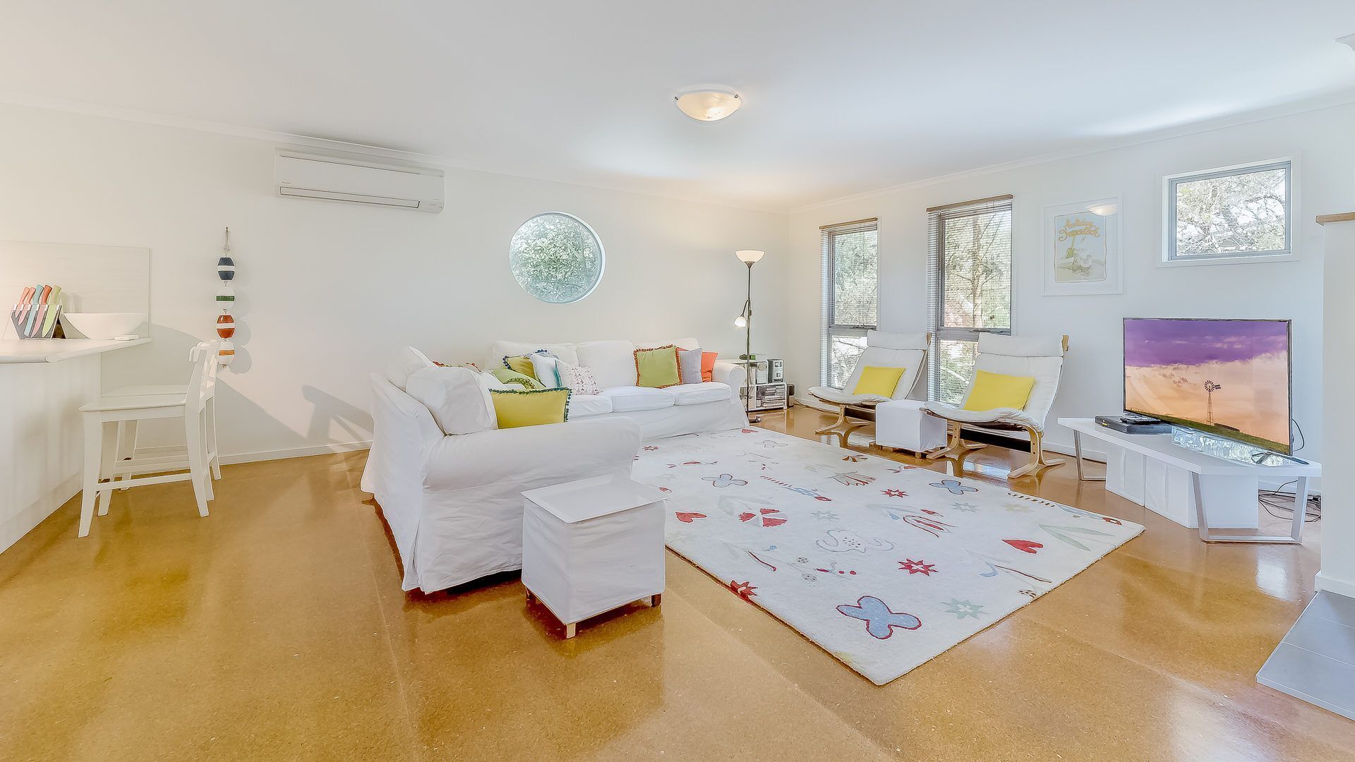 A modern living room with a white sofa, a coffee table, and a TV. There are two chairs and a small table. The room has large windows and a ceiling light.