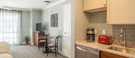 A compact kitchen with a dishwasher, a coffee maker, and a toaster. There is a dining area with a table and chairs, and a television in the background.