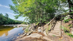 Eine hölzerne Treppe führt zu einem Flussufer, umgeben von Bäumen und Steinen.