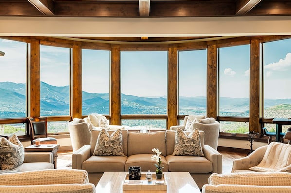 A spacious living area with a large window offering a mountain view, a beige sofa with patterned pillows, and a coffee table with a vase and a bottle.