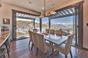 Uma sala de jantar com uma mesa grande e cadeiras, vista para montanhas através de portas de vidro e teto com luminárias pendentes.