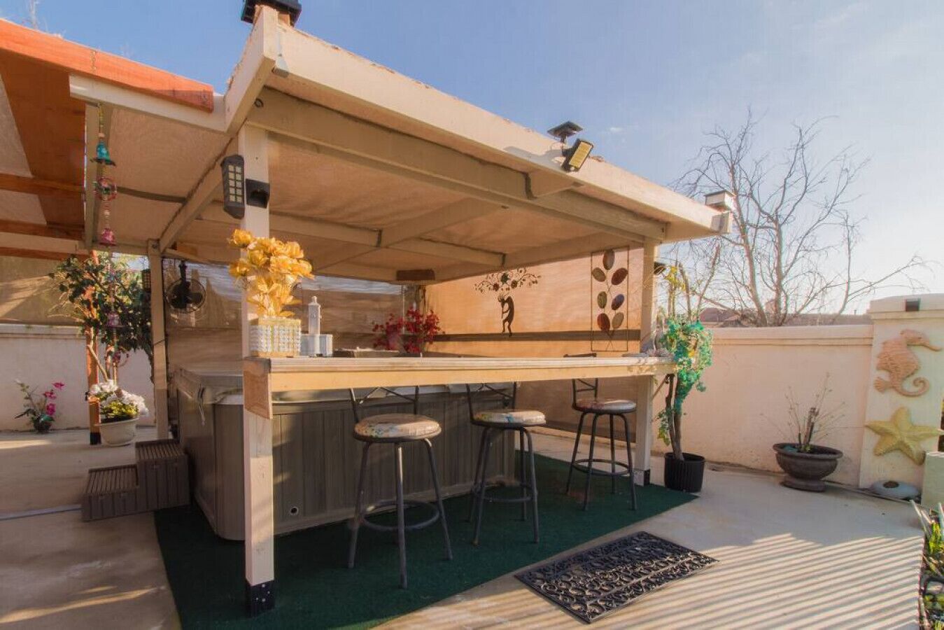 Un espace bar extérieur doté d’un comptoir en bois, de trois tabourets et d’un mur décoratif orné de motifs de feuilles.