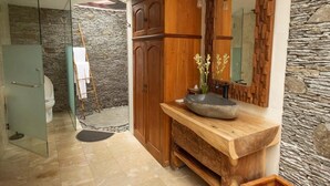 A bathroom with a stone wall, wooden vanity, and a glass shower enclosure. - Ubud Serendipity Villa (Ubud)