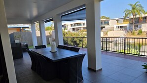 Een overdekt terras met een grote tafel en stoelen, een decoratief middelpunt en uitzicht op de omliggende gebouwen en een waterpartij.