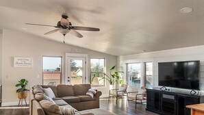 Ein geräumiges Wohnzimmer mit einer großen Eckcouch, einem Deckenventilator, einem Flachbildfernseher und Blick ins Freie.