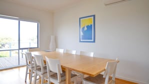 Un área de comedor con una mesa de madera y sillas blancas, un ventanal grande y un cuadro en la pared.
