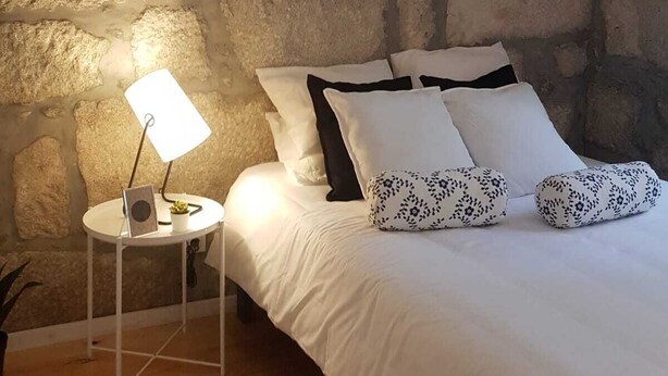 Un dormitorio con pared de piedra, una mesita de noche redonda con lámpara, y una cama con ropa de cama blanca y almohadas estampadas.