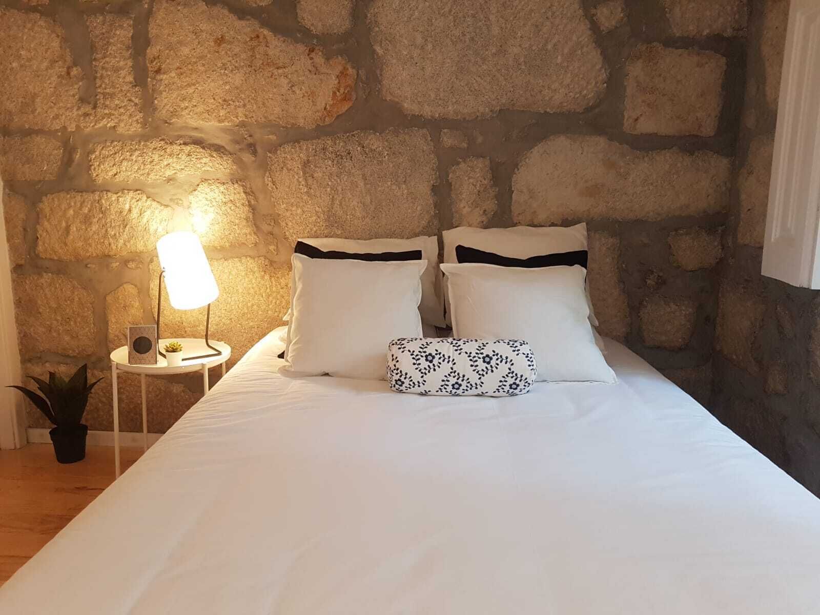 Un dormitorio con pared de piedra, una cama con ropa blanca y almohadas negras, una lámpara de noche y una planta pequeña.